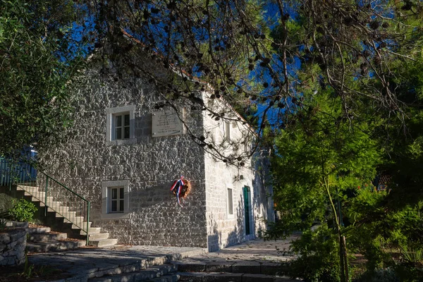 NECUJAM, CROATIA, 09.01.2025: Bir taş bina duvarında kırmızı, beyaz ve mavi kurdeleli bir çelenk ile bir hatıra plaketi barındırır. Güneşli adımlar ve derin mavi bir gökyüzü sakin tarihi bir sokak sahnesini canlandırıyor..