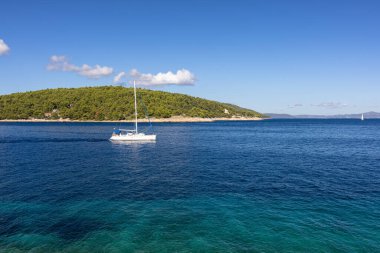 Derin mavi sularda süzülen sakin bir yelkenli, kayalık bir ön plan, ve parlak, berrak gökyüzü altında yeşil bir ada seyahat, doğa ve rahatlama temaları için mükemmel