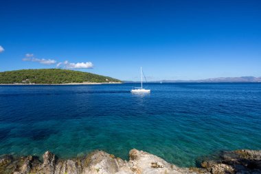 Derin mavi sularda süzülen sakin bir yelkenli, kayalık bir ön plan, ve parlak, berrak gökyüzü altında yeşil bir ada seyahat, doğa ve rahatlama temaları için mükemmel