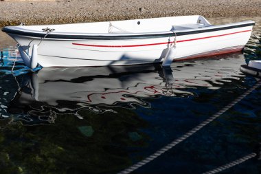 Sahil şeridinde beyaz bir tekne duruyor, kırmızı şeridi durgun suda yansıyor. Halatlar ve çamurluklar gövdede asılı, sakin bir sahil manzarası yaratıyor seyahat ve eğlence teması için mükemmel..