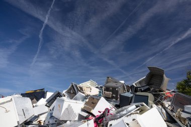 Çöpe atılmış bir yığın eşya, mobilya ve metal yığını açık bir hurdalıkta, parlak bir gökyüzüne karşı uzanıyor. Kaotik yığın atıkları, geri dönüşümü ve çevresel kaygıları vurguluyor.