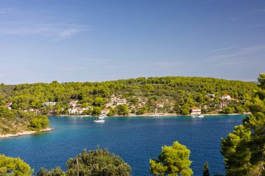 Çam örtülü bir manzara, sahil boyunca beyaz evler ve berrak mavi sulara demirlemiş birkaç yelkenli sahnesi. Seyahat, doğa ve eğlence temaları için ideal.