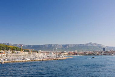 BÖLÜM, CROATIA, 09.06.2025: Teknelerin ve etrafındaki orman tepelerinin yer aldığı bir sahil şehrinin arka planına karşı Marina.
