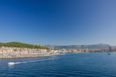 BÖLÜM, CROATIA, 09.06.2025: Teknelerin ve etrafındaki orman tepelerinin yer aldığı bir sahil şehrinin arka planına karşı Marina.