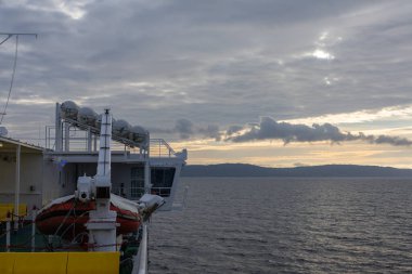 Gün batımında sakin denizleri aşan bir devriye gemisi cankurtaran botu, kurtarma ekipmanları ve güverte yapılarını gösteriyor. Uzak kara parçası ufku oluştururken dramatik bulutlar ışığı dağıtır..