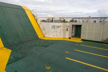 Kalın yeşil ve sarı güvenlik işaretleri olan geniş bir gemi güvertesi, merdivenler, kapı aralığı ve parmaklıklar. Korunmasız donanım ve yıpranmış yüzeyler engebeli deniz işleri ve endüstriyel faaliyet taşır.