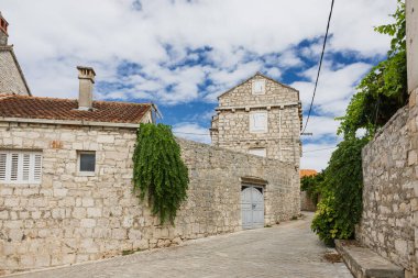 İçinde kaldırım taşı yolu, sarmaşık örtülü duvarlar, ağır ahşap bir kapı ve kepenkleri olan sessiz, ortaçağ tarzı taştan bir köy sokağı. Parlak gökyüzü, zamansız kırsal çekiciliği ve tarihi geliştirir..