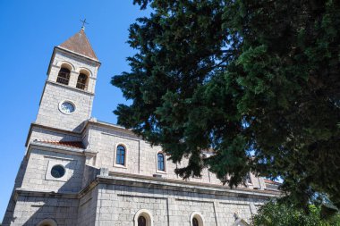 Tarihi bir taş kilisede uzun bir saat kulesi, kemerli pencereler ve fayanslı bir çatı bulunur. Mimari, gelenek ve barışçıl çevrenin altını çizen yemyeşil bir ağacı olan açık mavi gökyüzüne karşı..