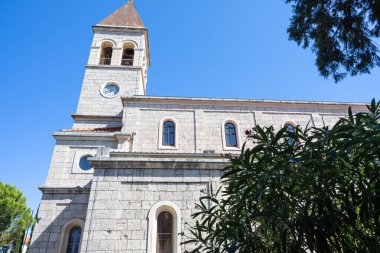 Tarihi bir taş kilisede uzun bir saat kulesi, kemerli pencereler ve fayanslı bir çatı bulunur. Mimari, gelenek ve barışçıl çevrenin altını çizen yemyeşil bir ağacı olan açık mavi gökyüzüne karşı..