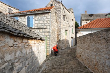 Parlak kırmızı bir scooter, tarihi bir köyde taştan duvarlarla çevrili, güneşli bir cadde boyunca dinleniyor. Kırsal dokular, dar şeritler ve sıcak ışık resimli bir seyahat sahnesini çağrıştırıyor..