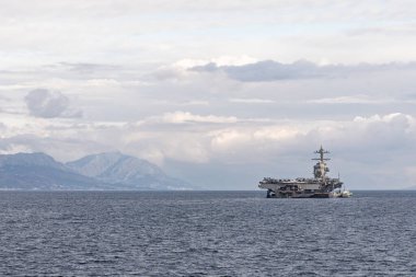 Muazzam bir uçak gemisi sakin sularda, engebeli bir kıyı şeridi ve gri bulutlu bir gökyüzünün altındaki uzak dağlarla yol alır. Ordu veya seyahat için uygun güçlü bir deniz manzarası.