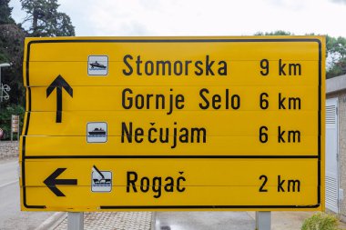 Parlak sarı bir yol tabelasında Stomorska, Gornje Selo ve Necujam da dahil olmak üzere çok sayıda Hırvat ada kasabasına uzaklıklar sıralanıyor. Sahne, güneşli bir açık havada seyahat, seyrüsefer ve turistik yol tabelasını öneriyor..