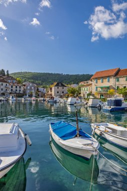 Sakin bir liman kenti boyunca, taştan evler, yemyeşil tepeler ve demirlemiş yelkenlilerin bulunduğu pitoresk bir sahil kasabası. Parlak gökyüzü ve berrak su huzurlu, kartpostala değer bir sahil manzarası yaratır..