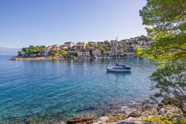 Suyun kenarında sakin bir liman köyü, rıhtımda tekneler ve yüzeyde dans eden güneş ışığı yansımaları. Büyüleyici evler ve yeşil tepeler huzurlu, manzaralı bir sahil havası yaratır..