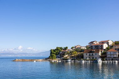 Sakin bir körfez boyunca sakin bir sahil köyü, küçük bir limana yanaşmış tekneler ve güneşli taş evler berrak mavi gökyüzünün altında yumuşak bir yamaca tırmanıyor..