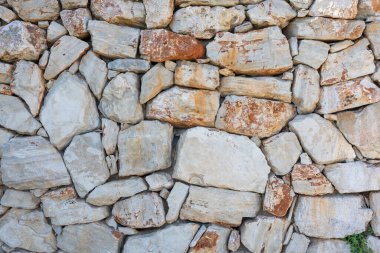 A weathered gray stone wall built from irregular rocks, showing layered masonry and hints of dry grass in the foreground. Ideal for rustic architecture, durability, and natural construction themes.