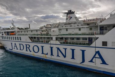 BÖLÜM, CROATIA, 10.25.2025: Bir Jadrolinija feribotu limanda beyaz gövdede koyu mavi harflerle oturuyor. Bulutlu gökyüzü, durgun su ve güverte aktiviteleri seyahat, seyahat ve deniz taşımacılığı titreşimlerini akla getiriyor...