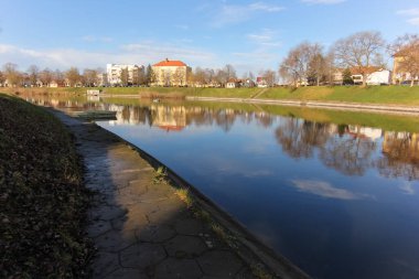Sakin nehir, açık gökyüzünün altında nehir boyunca uzanan patikalı binaları ve ağaçları yansıtıyor..