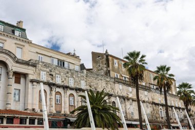 Yıpranmış bir taş bina, tarihi bir kıyı kasabasını andıran, palmiye kenarlı kıyı şeridi boyunca uzanır. Klasik mimari, kemerler ve kepenkli pencereler zamansız, seyahate hazır bir sahne yaratır..