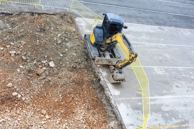Küçük çaplı bir kazıcı, parlak sarı güvenlik bariyerleriyle çevrili bir yol inşaatında siper kazıyor. Olay yeri kentsel gelişim, ağır ekipman kullanımı ve aktif site faaliyetlerini aktarıyor.