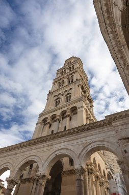 Kavisli bir taştan kilise cephesinin yanında, kemerler, kolonlar ve karmaşık oymalar bulunan yüksek bir çan kulesi yükseliyor. Gökyüzü yumuşak bulutlarla dolu, dramatik, ortaçağdan esinlenilmiş mimari bir sahne yaratıyor..