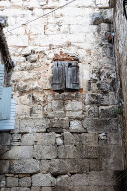 Küçük bir pencerenin üzerinde aşınmış ahşap bir kepenk olan engebeli taştan bir dış görünüş. Eskimiş dokular, yosun ve açığa çıkmış kablolar tarihi bir şehir ortamında eski, sessiz bir ara sokak havası yaratır..