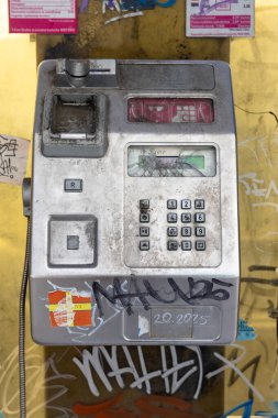 SPLIT, CROATIA, 10.25.2025: Yıpranmış bir ankesörlü telefon, afişler ve renkli etiketlerle çevrili, grafiti kaplı sarı bir duvarın karşısında oturuyor. Retro cihaz, sarmalanmış kablo, ve yontulmuş metal cesur şehir hayatı estetiğini ortaya çıkarıyor..