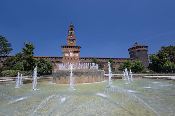 A sunlit European castle features a tall tower beside a circular fountain with jets. Lush trees and brick walls frame a timeless, cultural city scene under a clear blue sky.