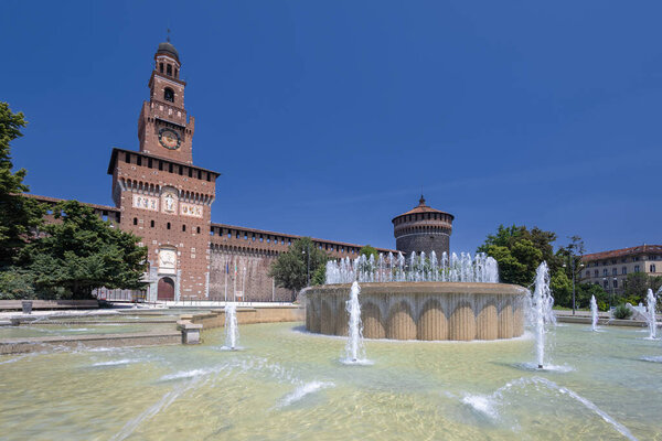 A sunlit European castle features a tall tower beside a circular fountain with jets. Lush trees and brick walls frame a timeless, cultural city scene under a clear blue sky.