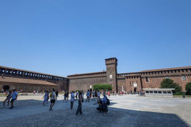 MILAN, ITALY, 06.19.2025: tarihi bir kalede parlak bir gün yüksek tuğlalı duvarlar, önemli bir ortaçağ kulesi ve turistlerle dolu geniş bir avlu. Açık meydan ve korunmuş mimari miras ve seyahati çağrıştırıyor.