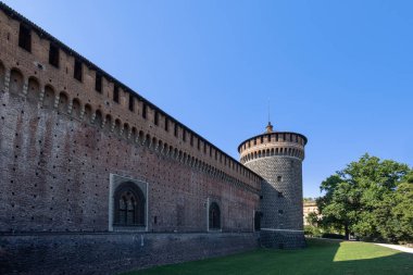 MILAN, İtalya, 06.19.2025: kemerli açıkları ve parlak mavi gökyüzünün altındaki yeşil çimlerin üzerinde yükselen uzun yuvarlak kulesi olan tarihi bir tuğla kale duvarı. Miras, mimari ve açık hava kale temaları için ideal..