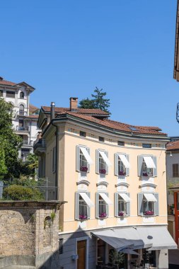 Beyaz kepenkleri, çiçek kutuları ve dekoratif kemerleri olan güneşli bir pastel ev. Klasik Avrupa mimarisi, kıvrımlı cephe, kiremitli çatı ve parlak mavi gökyüzü sıcak Akdeniz cazibesini çağrıştırıyor..