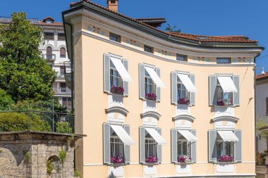 Beyaz kepenkleri, çiçek kutuları ve dekoratif kemerleri olan güneşli bir pastel ev. Klasik Avrupa mimarisi, kıvrımlı cephe, kiremitli çatı ve parlak mavi gökyüzü sıcak Akdeniz cazibesini çağrıştırıyor..