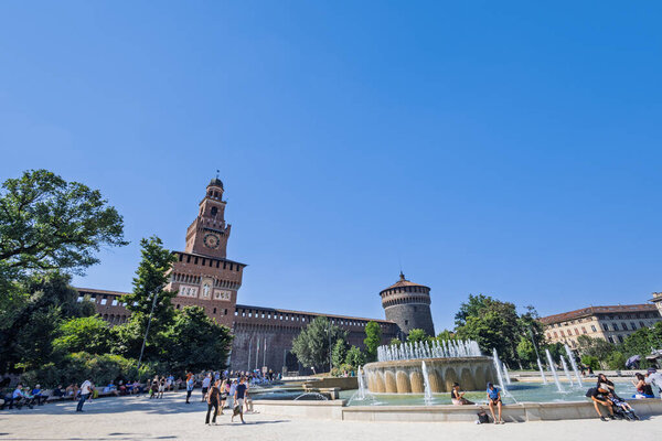 MILAN, ITALY, 06.19.2025:A sunlit European castle features a tall tower beside a circular fountain with jets. Lush trees and brick walls frame a timeless, cultural city scene under a clear blue sky.