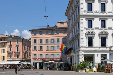 LUGANO, SWitness ZERLAND, 06.20.2025: Soluk pembe bir bina, büyük beyaz bir faade, açık kafeler, yayalar ve dikkat çekici bir gökkuşağı bayrağı içeren güneşli bir Avrupa şehir meydanı