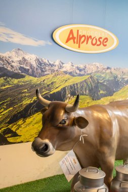 CASLANO, SWitZERLAND, 06.20.2025: Bronz inek heykeli yeşil tepeler ve kar tepeleri içeren canlı bir Alprose çikolata tabelasının altında duruyor. Süt ve şeker temaları için mükemmel, eğlenceli, kırsal bir ekran.