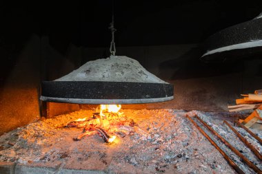 Büyük bir dökme demir kapağı parlayan bir demir ocağının üzerinde, dökmeye hazır bir kum kalıbı ile asılı duruyor. Alevler dumanlı bir atölyeyi aydınlatırken, odun ve aletler yakınlarda uzanıyor, taşımacılık, ısı ve endüstriyel beceri..