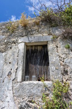 Kırsal bir taş duvar, üst ve yan taraflarında sarmaşık ve bitkiler büyüyen yıpranmış ahşap bir pencere çerçeveler. İki cam bardak güneşli bir çıkıntıda dinleniyor. Terk edilmişliği ve sessiz tarihi çağrıştırıyor..