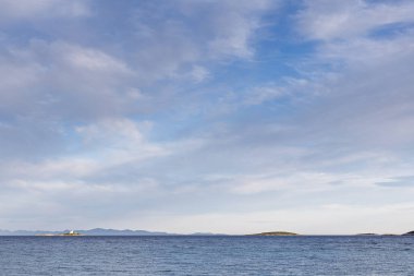 Sakin bir deniz, uzak adalar ve geniş mavi gökyüzünün altında soluk bir ufuk içeren sakin bir deniz manzarası. Seyahat, rahatlama ve doğadan esinlenilmiş açık hava görselleri için mükemmel..