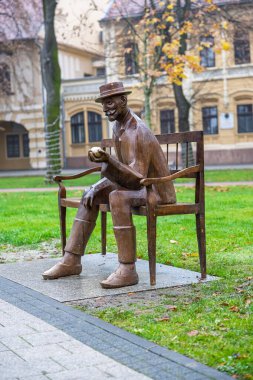 VINKOVCI, CROATIA, 12.13.2025: Şapka takan bir adamın bronz heykeli şehir parkındaki bir bankta, elinde elma tutarken oturmaktadır. Sonbahar yaprakları, ağaçlar ve tarihi bir bina sakin ve yansıtıcı bir zemin oluşturur..