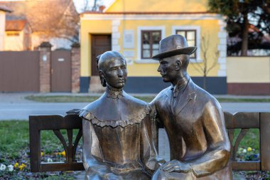 VINKOVCI, CROATIA, 12.27.2025: Parktaki bir bankta oturan bir erkek ve kadının bronz heykeli, açık sarı bir sokak zeminine karşı. Bir sanat sahnesinde romantizmi, arkadaşlığı ve zamansız şehir hayatını ifade eder..