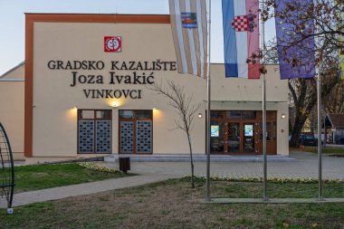 VINKOVCI, CROATIA, 12.27.2025: Hırvatistan 'ın Vinkovci kentindeki Şehir Tiyatrosu Joza Ivakiç' in dış görünüşü. Büyük bir tabela, dekoratif panelleri olan çift kapılar, çevre kaldırımlar ve bir çiçek bahçesi kültürel bir dönüm noktası oluşturur..