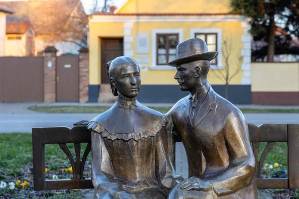 VINKOVCI, CROATIA, 12.27.2025: Parktaki bir bankta oturan bir erkek ve kadının bronz heykeli, açık sarı bir sokak zeminine karşı. Bir sanat sahnesinde romantizmi, arkadaşlığı ve zamansız şehir hayatını ifade eder..
