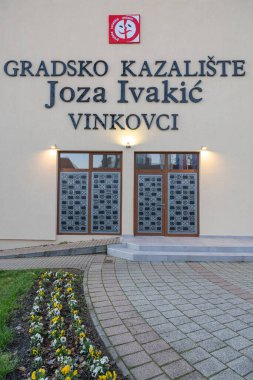 VINKOVCI, CROATIA, 12.27.2025: Hırvatistan 'ın Vinkovci kentindeki Şehir Tiyatrosu Joza Ivakiç' in dış görünüşü. Büyük bir tabela, dekoratif panelleri olan çift kapılar, çevre kaldırımlar ve bir çiçek bahçesi kültürel bir dönüm noktası oluşturur..