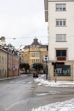 CELJE, SLOVENIA, 01.03.2026: Süslü camları, modern vitrinleri, park edilmiş bir arabası ve kaldırım boyunca kar bulunan sakin, atmosferik bir şehir havası yaratan tarihi sarı bir binanın yer aldığı sakin bir şehir sokak manzarası.