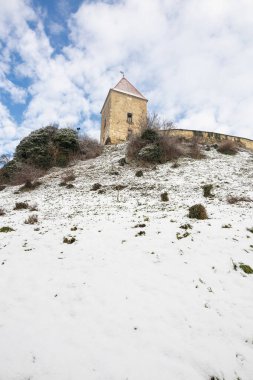 Bir taş kule karla kaplı bir tepenin üzerinde, seyrek çalılıklar ve yıpranmış bir duvarla çevrili. Açık mavi gökyüzü, kışın sakin ve tarihi bir kaleyi vurguluyor..