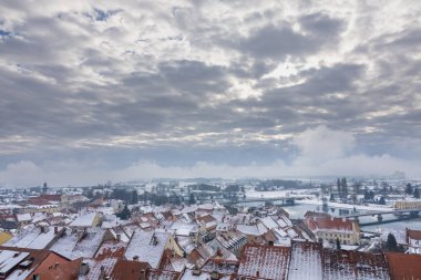 Sakin bir nehir boyunca karla kaplı bir kasabanın sakin bir kış manzarası, sisli, bulutlu bir gökyüzünün altında kemerli bir köprü ve renkli çatılar var..