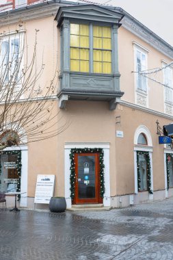 PTUJ, SLOVENIA, 01.04.2026: Büyüleyici bir bej bina, neşeli bir çelenkle çerçevelenmiş bir kapının üzerinde gri süslemeli bir pencere balkonuna sahiptir. Karlı, kaldırımlı sokak manzarası kış turizmini çağrıştırıyor.
