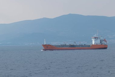 Turuncu kimyasal tanker gemisi, deniz taşımacılığı, kargo lojistiği ve sanayi taşımacılığını gösteren dağlık bir kıyı şeridi yakınındaki sakin mavi sular boyunca ilerlerken, hafif ışık altında yalnızlık, seyahat ve küresel ticareti anımsatıyor..