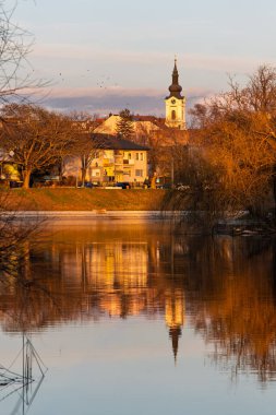 Sakin nehir suyuna yansıyan, dinginlik ve nostalji uyandıran, sıcak ışık, yapraksız ağaçlar ve binalarla yıkanmış tarihi bir kilise saat kulesinin altın saatinde huzurlu küçük bir kasaba sahnesi..